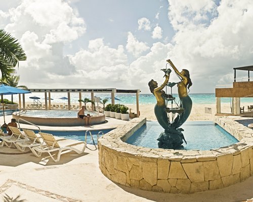 An outdoor swimming pool with hot tub and chaise lounge chairs and sunshades alongside the sea.