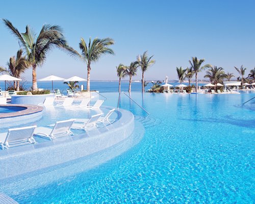 An outdoor swimming pool with chaise lounge chairs and sunshades alongside the ocean.