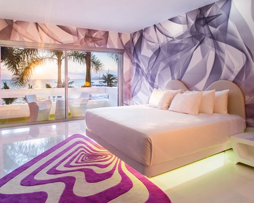 A well furnished bedroom with a balcony and view of palm trees alongside the ocean at dusk.