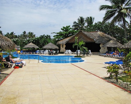 An outdoor swimming pool with patio furniture chaise lounge chairs and thatched sunshade.