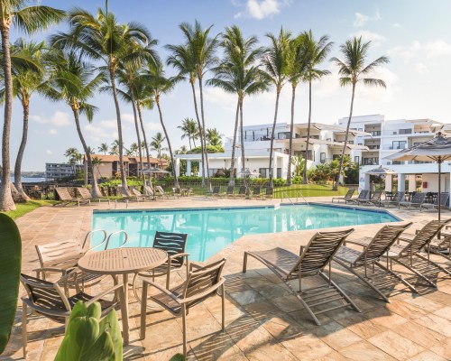 AN OUTDOOR SWIMMING POOL WITH CHAISE LOUNGE CHAIRS AND PATIO FURNITURE ALONGSIDE MULTI STORY RESORT UNITS.