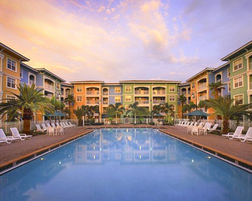 An outdoor swimming pool with chaise lounge chairs alongside multi story resort units at dusk.