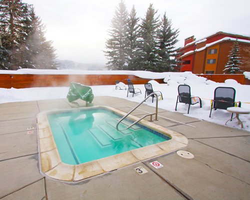 Outdoor hot tub with patio chairs covered in the snow alongside the wooded area.