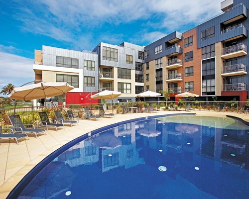 An outdoor swimming pool with chaise lounge chairs and sunshades alongside multi story units.