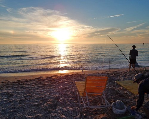 A man fishing in the beach with chaise lounge chairs at dusk.