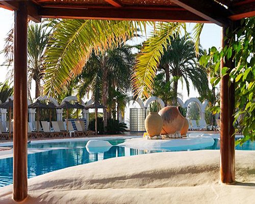 Outdoor swimming pool with chaise lounge chairs thatched sunshades and palm trees.