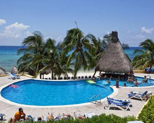 Thatched covered bar on outdoor swimming pool with chaise lounge chairs and palm trees alongside the beach.