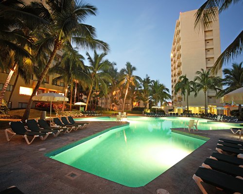 An outdoor swimming pool with chaise lounge chairs alongside the resort at night.