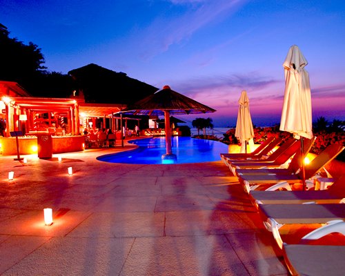An outdoor swimming pool with chaise lounge chairs and sunshades at night.