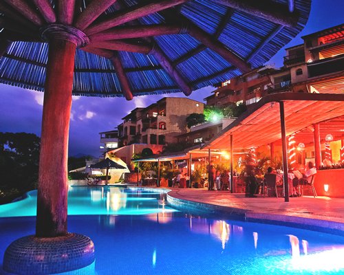An outdoor swimming pool with sunshades alongside multi story resort units at night.