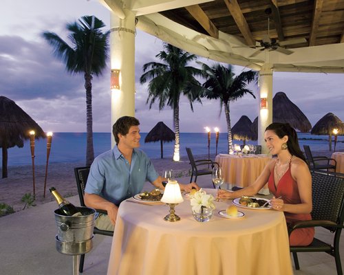 A couple in a dining area alongside the beach with thatched sunshades.