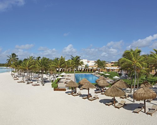 An outdoor swimming pool with chaise lounge chairs and thatched sunshades alongside the beach.