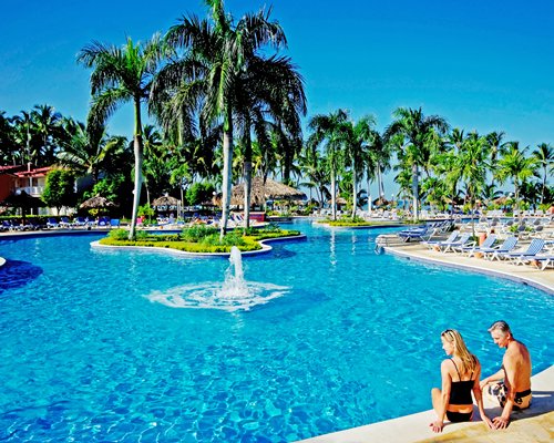 A couple in the outdoor swimming pool with the water fountain and chaise lounge chairs surrounded by trees.