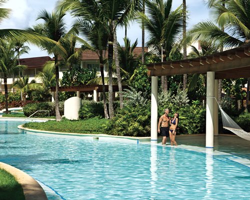 An outdoor swimming pool with chaise lounge chairs and thatched covered bar surrounded by trees.