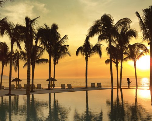 An outdoor pool with chaise lounge chairs and coconut trees alongside the beach at dusk.