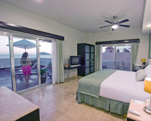 A well furnished bedroom with a woman standing at the balcony with chaise lounge chairs sunshade and ocean view.