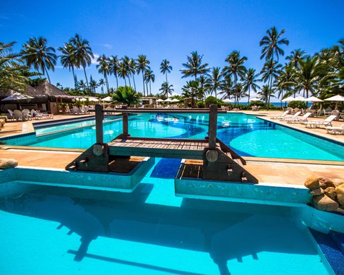 Large outdoor swimming pool with a bridge chaise lounge chairs and sunshades surrounded by palm trees.