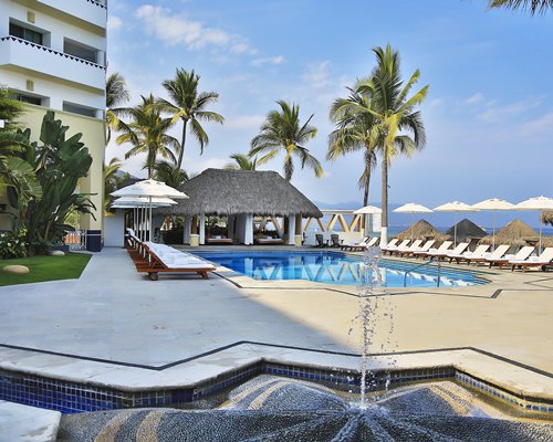 An outdoor swimming pool with chaise lounge chairs sunshades and a water fountain alongside resort units.