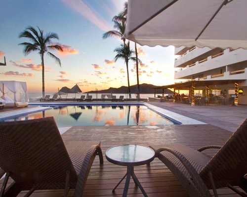 An outdoor swimming pool with chaise lounge chairs alongside multi story resort units at dusk.