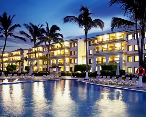 An outdoor swimming pool with chaise lounge chairs alongside the resort at night.