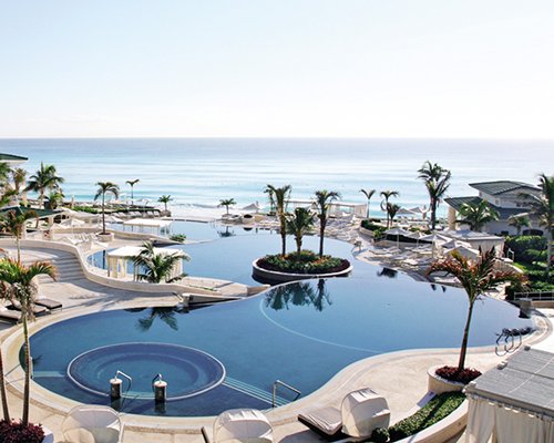Large outdoor swimming pool with chaise lounge chairs sunshades and palm trees alongside the sea.