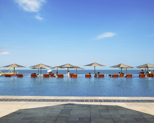 An outdoor swimming pool with chaise lounge chairs and sunshades alongside the sea.