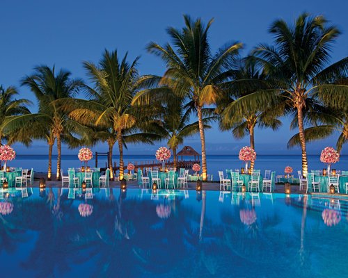 A large outdoor swimming pool with chaise lounge chairs sunshades and trees alongside the ocean.