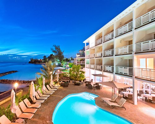 An outdoor swimming pool with chaise lounge chairs alongside resort units and the ocean.