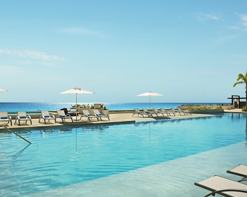 An outdoor swimming pool with chaise lounge chairs and sunshades alongside the ocean.