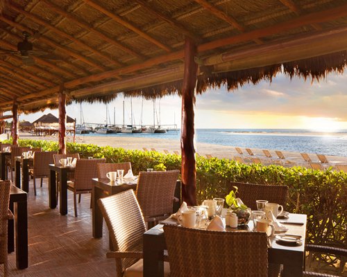 Thatched covered outdoor restaurant alongside the beach with chaise lounge chairs and boats.