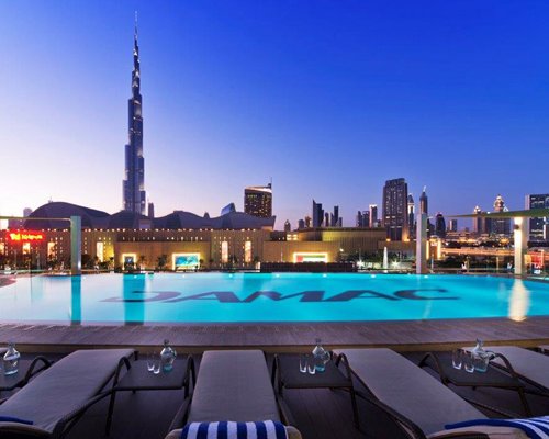 Exterior view of buildings and Damac Maison Dubai Mall Street with outdoor swimming pool and chaise lounge chairs.
