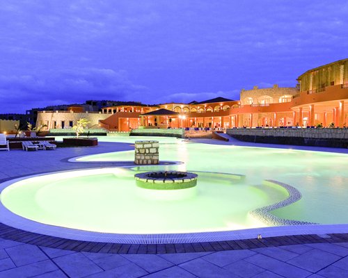 An outdoor swimming pool with chaise lounge chairs alongside multi story resort units at dusk.