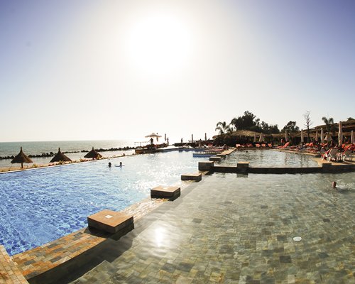 An outdoor swimming pool with thatched sunshades and chaise lounge chairs alongside the ocean.