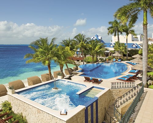 An outdoor swimming pool with a hot tub chaise lounge chairs and sunshades alongside the ocean.