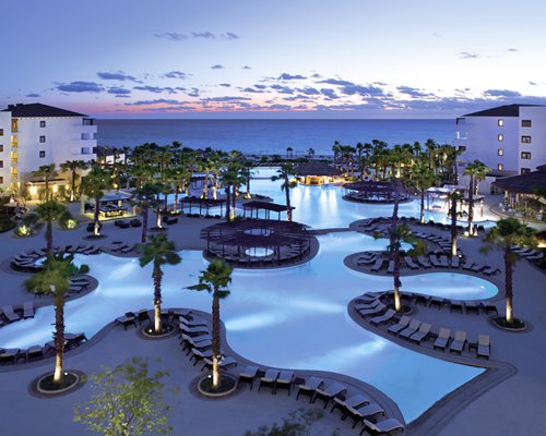 Large outdoor swimming pool with chaise lounge chairs and palm trees alongside the sea.