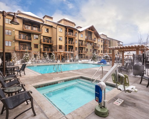 AN OUTDOOR SWIMMING POOL WITH HOT TUB AND CHAISE LOUNGE CHAIRS ALONGSIDE THE RESORT COVERED IN SNOW.