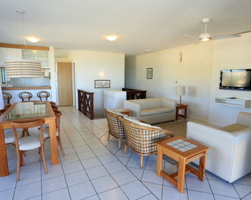 A WELL FURNISHED LIVING ROOM WITH A TELEVISION AN OPEN PLAN KITCHEN AND DINING AREA.