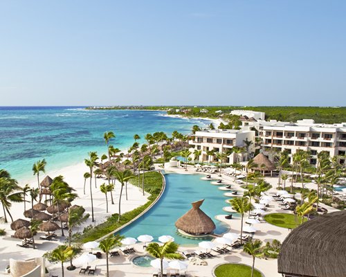 An outdoor swimming pool with trees thatched sunshades and chaise lounge chairs alongside the sea.