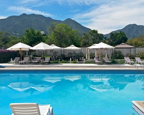 An outdoor swimming pool with chaise lounge chairs and sunshades surrounded by trees alongside the mountain.