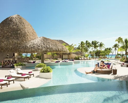 An outdoor swimming pool with thatched sunshades chaise lounge chairs and trees.