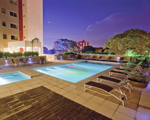 An outdoor swimming pool with chaise lounge chairs alongside the resort at night.