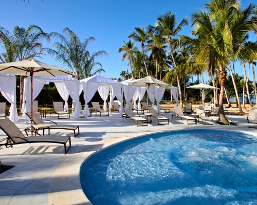 OUTDOOR SWIMMING POOL WITH CHAISE LOUNGE CHAIRS SUNSHADES AND PALM TREES.