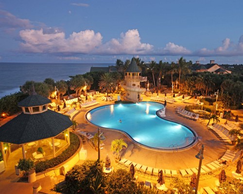 AN AERIAL VIEW OF THE WELL LIT OUTDOOR SWIMMING POOL ALONGSIDE THE OCEAN.