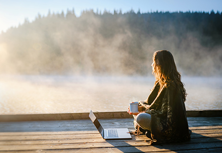 A woman sitting on the dock of a misty body of water with a mug of coffee and her laptop